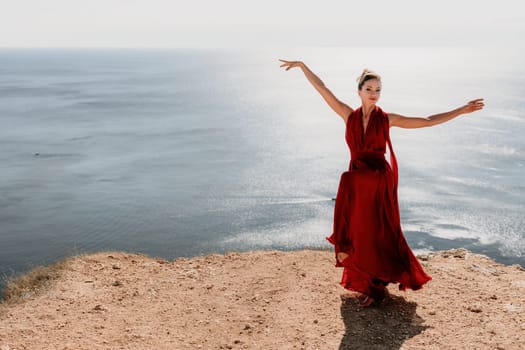 Side view a Young beautiful sensual woman in a red long dress posing on a rock high above the sea during sunrise. Girl on the nature on blue sky background. Fashion photo.