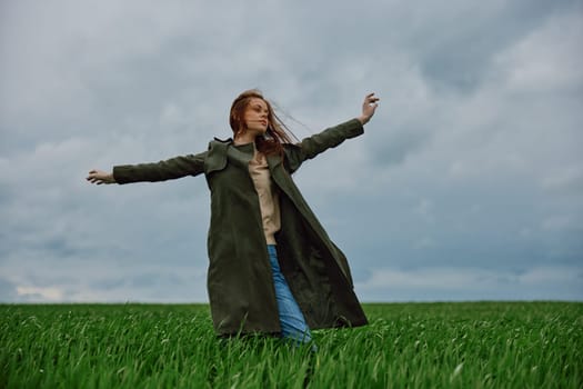 a woman in a long coat stands in a field against a cloudy sky with her back to the wind with her hands raised behind her head. High quality photo