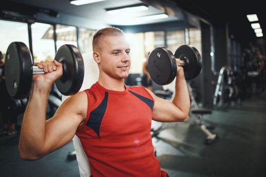 Shot of a muscular guy in sportswear working out at the hard training in the gym. He is pumping his shoulder muscle with dumbbell.