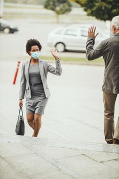 Shot of successful black businesswoman and her senior male colleague using protective mask and greeting with wave hands during COVID-19 pandemic to avoid handshakes.