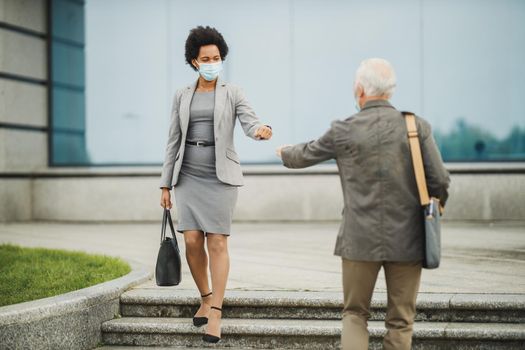 Shot of successful black businesswoman and her senior male colleague using protective mask and greeting with bumping fists outdoors during COVID-19 pandemic to avoid handshakes.