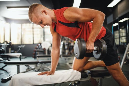 Shot of a muscular guy in sportswear working out at the hard training in the gym. He is pumping his back muscle with dumbbell.