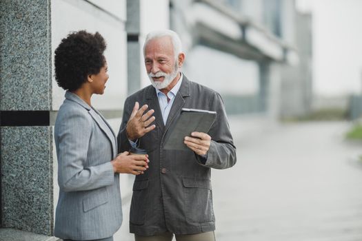 Shot of successful senior businessman and his black female colleague talking and using digital tablet during a coffee break in front of the office building.
