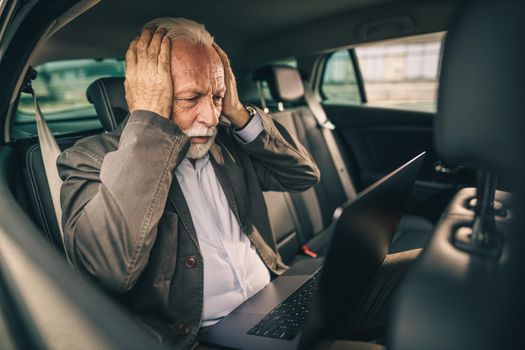Shot of a stressed senior man using laptop while sitting in the backseat of a car during his business commute.