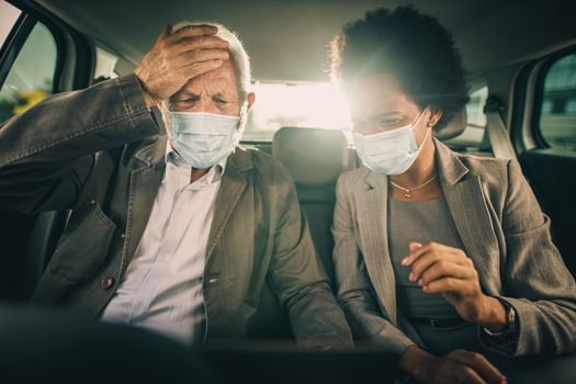 Shot of two worried multi-ethnic people with protective mask using laptop and having a discussion while sitting in the backseat of a car on their business commute during COVID-19 pandemic.