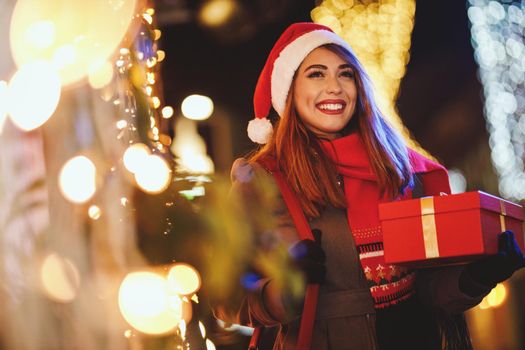 Shoot of a cheerful young woman with red present box is having fun in the city street at Christmas time.