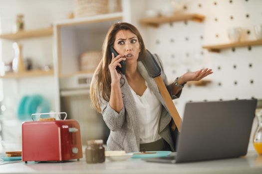 Shot of a multi-tasking young business woman talking on her smartphone and using laptop in her kitchen while getting ready to go to work.