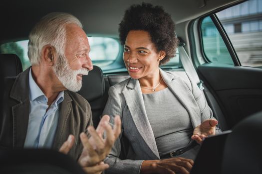 Shot of two successful multi-ethnic people using laptop and having a discussion while sitting in the backseat of a car during their business commute.