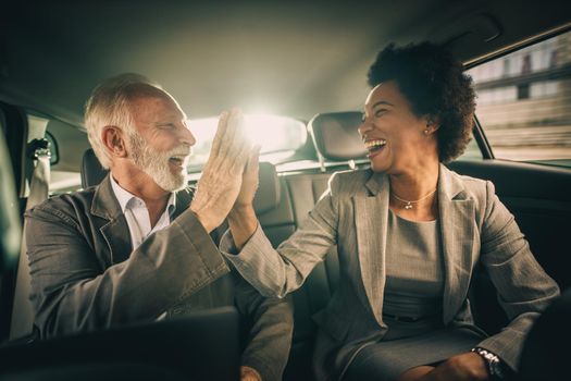 Shot of two successful multi-ethnic people giving five and celebrate good work while sitting in the backseat of a car during their business commute.
