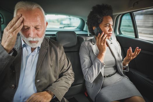 Shot of two successful multi-ethnic business people using smartphone and laptop while sitting in the backseat of a car during their morning commute.