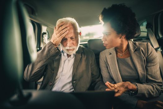 Shot of two worried multi-ethnic people using laptop and having a discussion while sitting in the backseat of a car during their business commute.