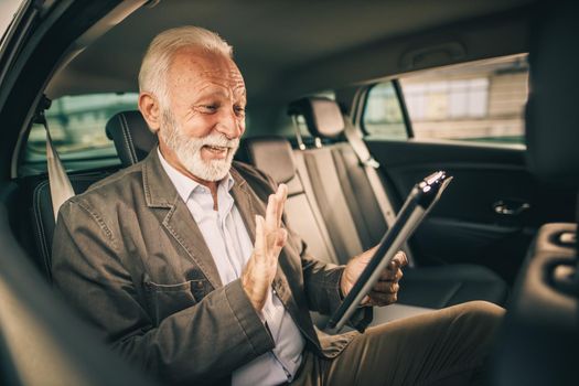 Shot of a successful senior man having video call on a digital tablet while sitting in the backseat of a car during his business commute.