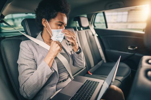 Shot of a successful black woman with protective mask working on laptop while sitting in the backseat of a car on her business commute during COVID-19 pandemic.