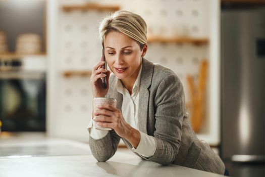 Shot of a mature woman drinking coffee and talking on smartphone in her kitchen while getting ready to go to work.