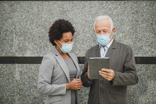 Shot of two successful multi-ethnic business people with protective mask using digital tablet and having a discussion while standing against a wall of corporate building during COVID-19 pandemic.