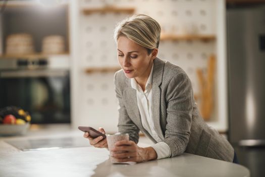 Shot of a mature woman drinking coffee and using smartphone in her kitchen while getting ready to go to work.