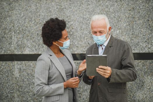 Shot of two successful multi-ethnic business people with protective mask using digital tablet and having a discussion while standing against a wall of corporate building during COVID-19 pandemic.