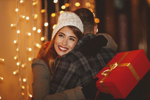 Shoot of a cheerful young couple are having fun in the city street at the Christmas night. They are hugging and giving holidays presents to each other.