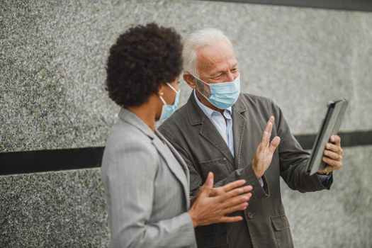 Shot of successful senior businessman and his black female colleague with protective mask using digital tablet and having a video call while standing against a wall of corporate building during COVID-19 pandemic.