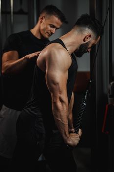 Shot of a muscular guy in sportswear working out with personal trainer at the gym. He is doing cable triceps pulldowns during a strength training.