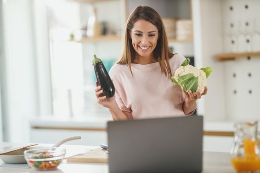 Shot of a young woman using laptop to make a vlog with someone while preparing a healthy meal at home.