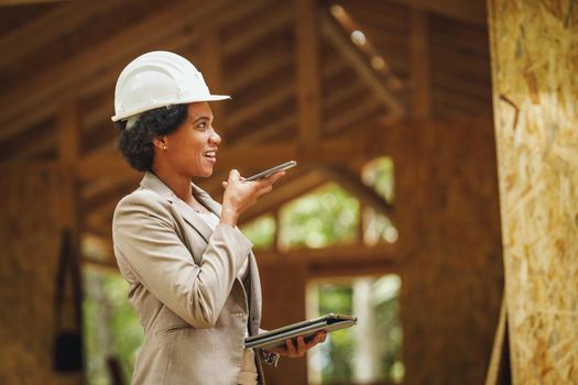 Shot of an African female architect with white helmet using a smartphone and checking construction site of a new wooden house.