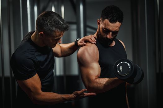 Shot of a muscular guy in sportswear working out with personal trainer at the gym. He is pumping up biceps muscule with heavy weight.