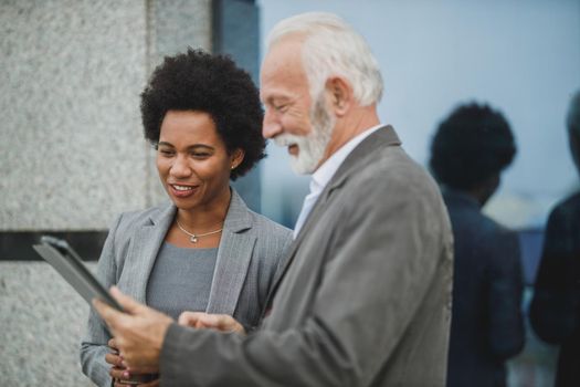 Shot of successful black businesswoman and her senior male colleague using digital tablet and having a discussion during a coffee break in front of the office building.