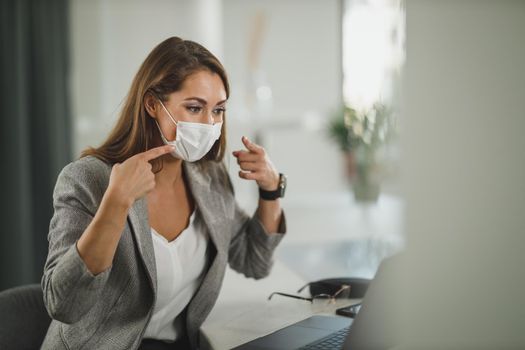 Shot of a young business woman with protective mask sitting alone in her office and having video call on laptop during corona virus pandemic.