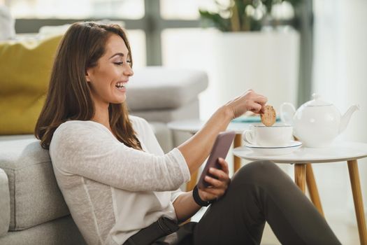 Beautiful, young woman enjoying tea time, holding a smart phone and surfing social media at her home.