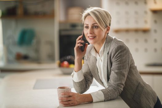 Shot of a mature woman drinking coffee and talking on smartphone in her kitchen.