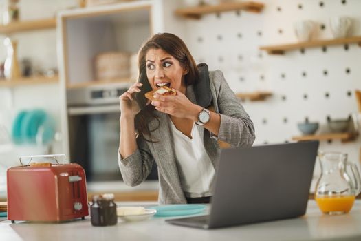 Shot of a multi-tasking young business woman having a breakfast and talking on smartphone in her kitchen while getting ready to go to work.