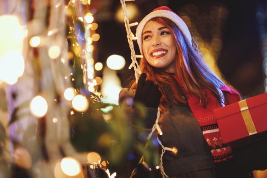 Shoot of a cheerful young woman with red present box is having fun in the city street at Christmas time.