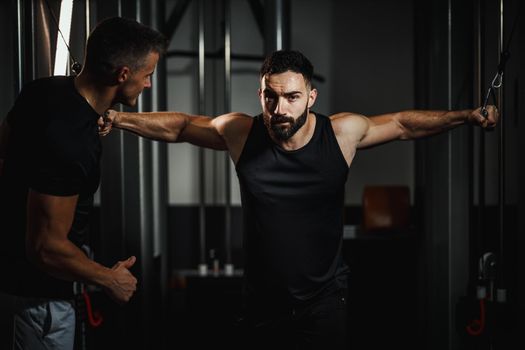 Shot of a muscular guy in sportswear working out with personal trainer at the gym. He is doing hard exercises for chest on cable machine during a strength training.