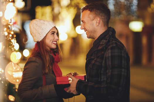 Shoot of a happy young couple are having fun in the city street at the Christmas night. Girl in love looking excited after receiving a Christmas gift from a her boyfriend.