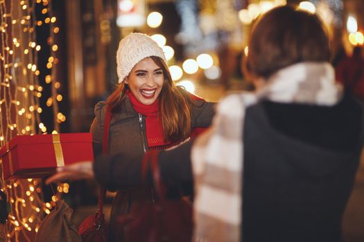 Cheerful two young women are having fun in the city street at the Christmas night. They are laughing and giving holidays presents to each other.