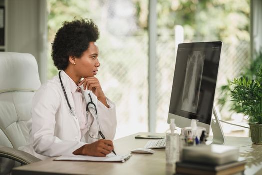 Shot of an African female doctor sitting alone in her consulting room and looking X-ray on computer during COVID-19 pandemic.