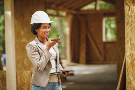 Shot of an African female architect with white helmet using a smartphone and checking construction site of a new wooden house.