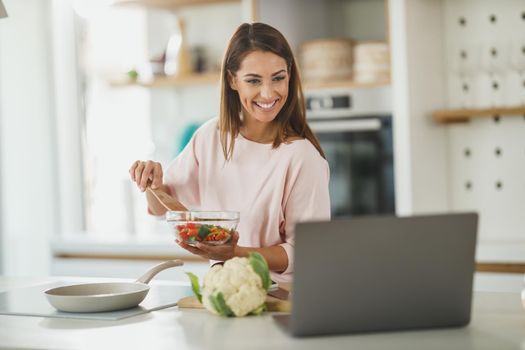 Shot of a young woman using a laptop while preparing a healthy meal at home.