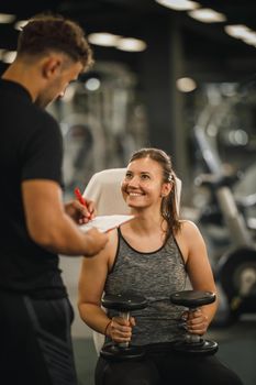 Shot of a muscular young woman in sportswear working out with personal trainer at the gym machine. She is pumping up her muscule with dumbbell.