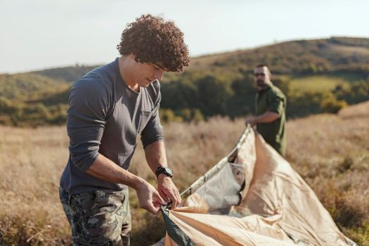 Group of a cheerful young friends putting up their tent on a suitable place in a meadow.