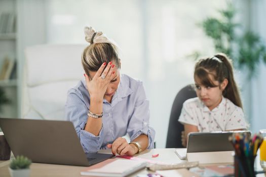 Overworked  single mother using laptop while her cute daughter learning at home.