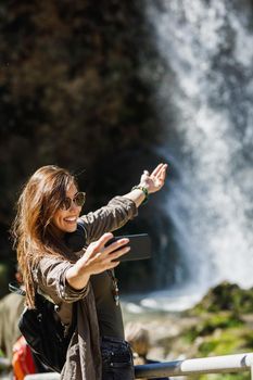 A smiling young woman making selfie with her smartphone while enjoying the view of the beautiful waterfall.