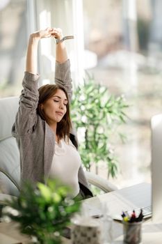 A young business woman looking relaxed with her hands over her head while sitting at her desk and working on computer.