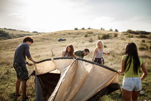 Group of a cheerful young friends putting up their tent on a suitable place in a meadow.