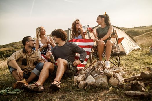 A group of cheerful friends sitting outside their tent at a festival and enjoying drink.