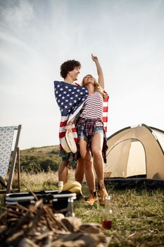 An attractive cheerful couple with american flag enjoying and partying in front a tent at the campsite.