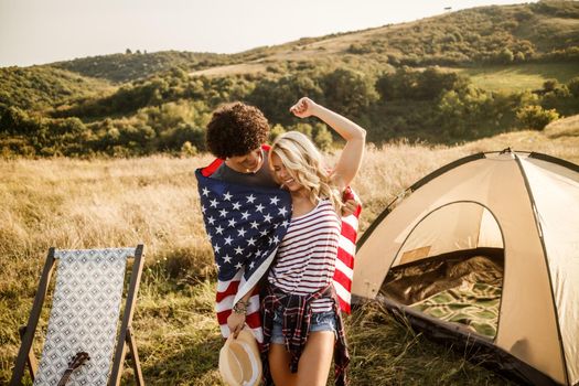 An attractive couple in love wrapped in american flag enjoying and partying in front a tent at the campsite.
