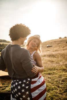 An attractive couple in love wrapped in american flag enjoying in front a tent at the campsite.