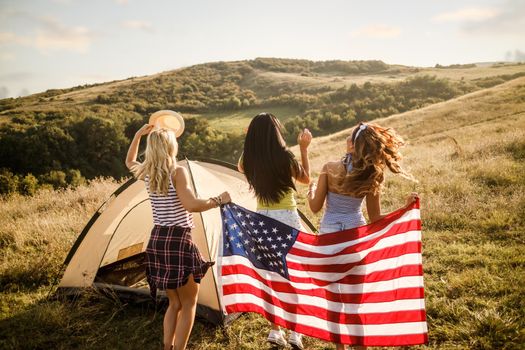 An attractive young female friends with american flag enjoying and partying in front a tent at the campsite.